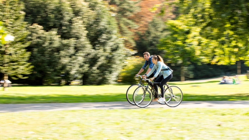 Zwei Radler:innen fahren durch den sonnigen Park, seitliche Ansicht